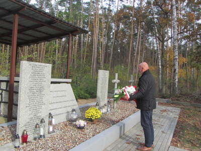 Zdjęcie przedstawia mogiłę zbiorową ogrodzoną płotem usytuowaną w lesie.