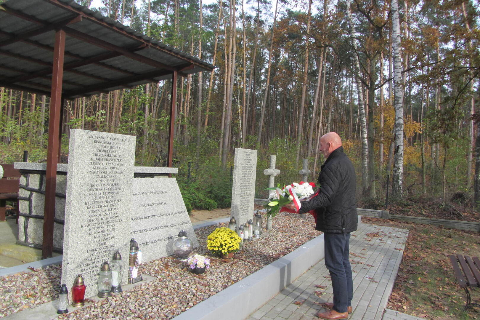 Zdjęcie przedstawia mogiłę zbiorową ogrodzoną płotem usytuowaną w lesie.