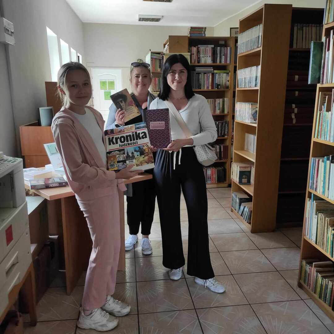 zdjęcie wykonane wewnątrz pomieszczenia. Na zdjęciu widoczne trzy osoby oglądające książki.
