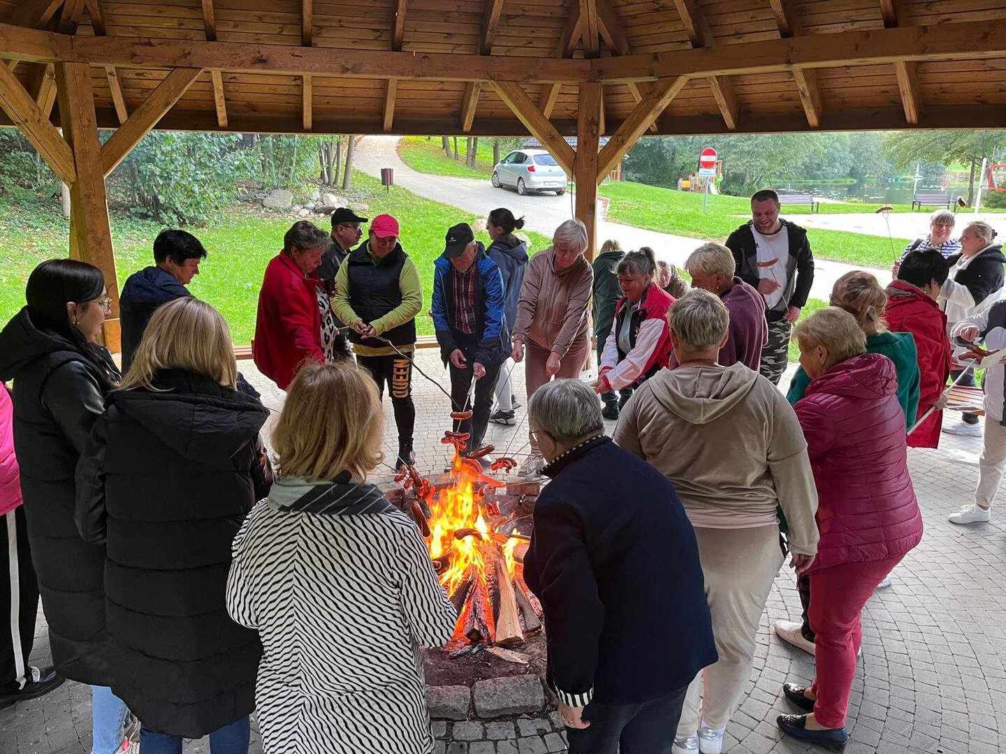 Grupa osób stojąca wokół palącego się ogniska i piekąca kiełbaski. Na zewnątrz.