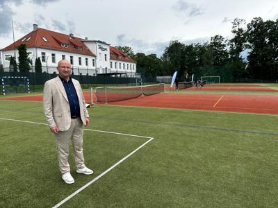 Zdjęcie przedstawia Wicestarostę w jasnym garniturze i ciemnej koszuli, stojącego na korcie tenisowym. 