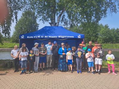 Grupa ludzi, w tym kilkoro dzieci, pozująca do zdjęcia przed niebieskim namiotem, obok zbiornika wodnego. Wiele dzieci trzyma trofea