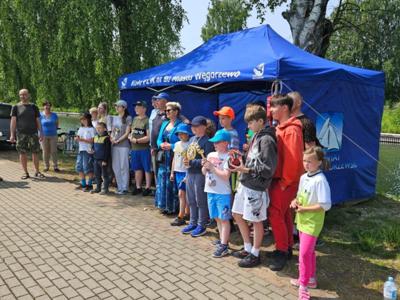 Szereg ludzi, w tym dorośli i dzieci, stojących na utwardzonej ścieżce obok zbiornika wodnego. 