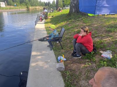 Osoba w żółtej czapce i koszulce z długim rękawem siedzi na czarnym składanym krześle na betonowym nabrzeżu, łowiąc ryby z wędką wyciągniętą nad wodę.