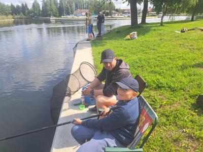 Mężczyzna w czarnej czapce i chłopak w granatowej czapce i okularach siedzą na brzegu kanału, łowiąc ryby.