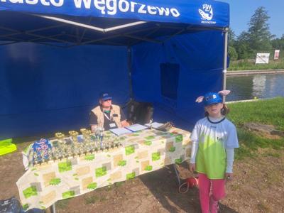 Młoda dziewczynka w różowych spodniach, biało-zielonej koszulce i niebieskiej czapce z uszami stoi przed stołem zastawionym trofeami. 