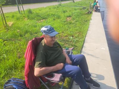 Starszy mężczyzna w zielonej koszulce i niebieskiej czapce siedzi na składanym krześle na trawiastym brzegu kanału, trzymając wędkę. 
