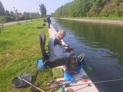 Mężczyzna w jasnej bluzie siedzi na składanym krześle na betonowym nabrzeżu, łowiąc ryby. 
