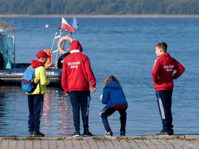 goście obserwują kajakarzy stojąc przy jeziorze, w tyle widać panoramę jeziora Mamry