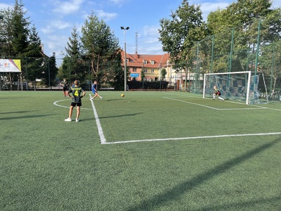 Zawodnik strzelający piłką w bramkę, za nim zawodnicy, w tle budynki