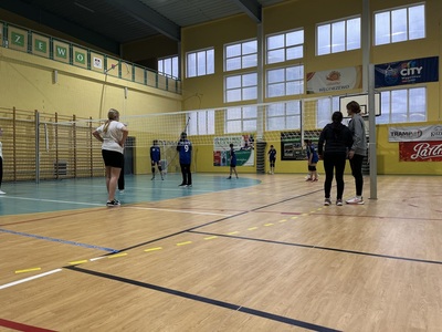 dwie drużyny rywalizują pod siatką na boisku obok stoją trenerki 