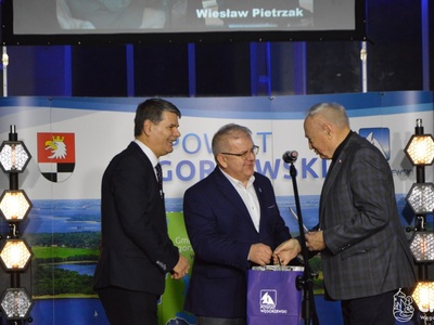 Burmistrz oraz Wicestarosta wręczają nagrodę dla pana Wiesława Pietrzaka. W tle baner powiatu węgorzewskiego.