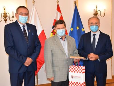 Zdjęcie pracownika Powiatu Kaliskiego. 
Na zdjęciu trzy osoby. Od strony lewej w garniturze koloru granatowego Wicestarosta Powiatu Kaliskiego Zbigniew Słodowy, druga osoba od strony lewej w marynarce koloru szarego Pan Zbigniew Witkowski, trzecia osoba od strony lewej w garniturze koloru czarnego Starosta Kaliski Krzysztof Nosal. Osoby wyposażone w maseczki stoją na tle trzech flag: flagi Rzeczpospolitej Polski, flagi Powiatu Kaliskiego oraz flagi Unii Europejskiej. Za flagami biała ściana.