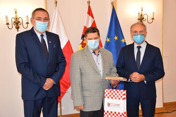 Zdjęcie pracownika Powiatu Kaliskiego. 
Na zdjęciu trzy osoby. Od strony lewej w garniturze koloru granatowego Wicestarosta Powiatu Kaliskiego Zbigniew Słodowy, druga osoba od strony lewej w marynarce koloru szarego Pan Zbigniew Witkowski, trzecia osoba od strony lewej w garniturze koloru czarnego Starosta Kaliski Krzysztof Nosal. Osoby wyposażone w maseczki stoją na tle trzech flag: flagi Rzeczpospolitej Polski, flagi Powiatu Kaliskiego oraz flagi Unii Europejskiej. Za flagami biała ściana. 