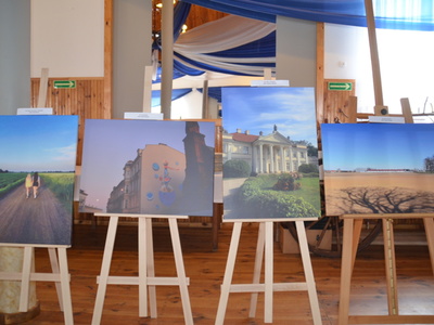 kolejne zdjęcia laureatów w dużym formacie 60 cm x 40 cm wystawione na sztalugach, od lewej dwie dziewczynki idące ścieżką wśród zielonych pól, obok zdjęcie budynku, którego ozdabia kolorowy mural przedstawiający bajkową postać wśród baniek mydlanych, trzecie zdjęcie przedstawia budynek Teatru im. Wojciecha Bogusławskiego w Kaliszu, czwarte zdjęcie od lewej to fotografia cienia rzuconego przez drzewo na tle zaoranego pola