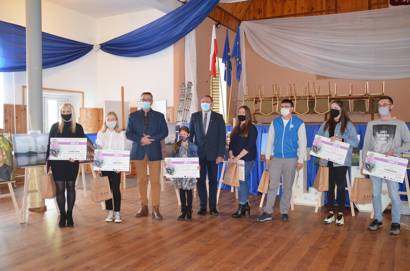 zdjęcia w sali OSP od lewej strony ubrana na czarno Justyna Karolak zdobywczyni III miejsca, następnie Julia Pietrzak zdobywczyni I miejsca, ubrana w bluzkę koloru białego oraz spodnie koloru czarnego, trzecia osoba od lewej Wójt Gminy Brzeziny Marek Cieślarczyk ubrany w marynarkę koloru granatowego oraz spodnie w kolorze musztardowym, Gabriela Kupajczyk oraz Prezes Zarządu Stowarzyszenia " Długosz Królewski" Zbigniew Słodowy wraz z laureatami konkursu. 