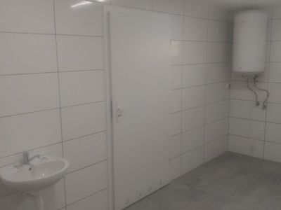 zdjęcie ściany wykonanej z białych płytek z połyskiem, na ścianie wisi umywalka w kolorze białym, obok drzwi w kolorze białym. W narożniku ściany widać biały zbiornik na wodę 