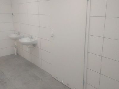 wygląd łazienki po remoncie, dwie białe umywalki wiszą na ścianie z białych płytek z połyskiem, na podłodze płytki w kolorze szarym 