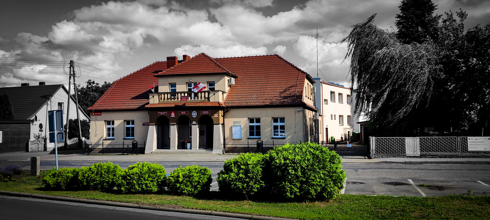 Na zdjęciu widać budynek urzędu gminy z czerwoną dachówką, flagą na balkonie, otoczony zielenią. W tle są inne budynki i pochylone drzewo, niebo jest częściowo pochmurne.