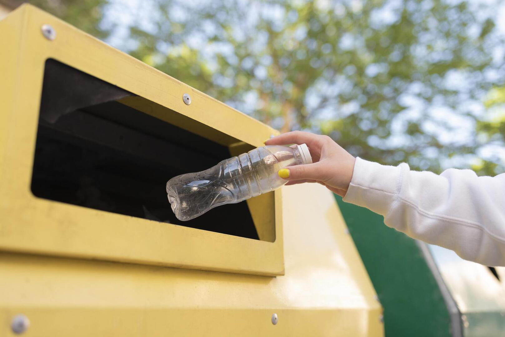 Zbliżenie dłoni osoby wkładającej zgniecioną plastikową butelkę do żółtego pojemnika na recykling. Osoba ma pomalowane na żółto paznokcie i nosi białą bluzę z długim rękawem. Tło przedstawia zieloną roślinność i błękitne niebo, sugerujące otoczenie na zewnątrz.