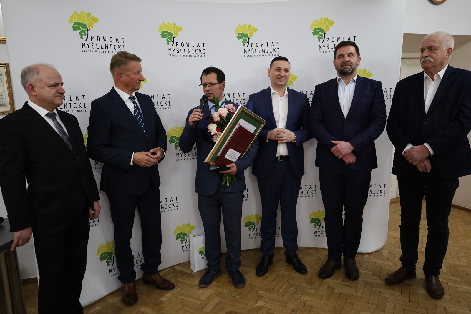 Widoczny laureat, w tym przedstawiciele Powiatu Myślenickiego, pozują wspólnie na tle ścianki z logo Powiatu Myślenickiego.