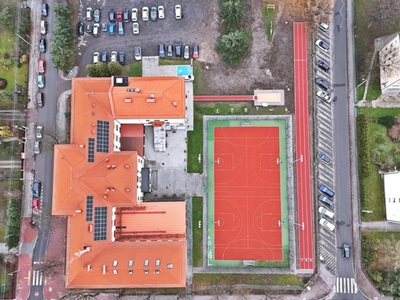 Widok z drona pokazujący dach szkoły, boisko sportowe, bieżnię, skocznię w dal i zaparkowane auta