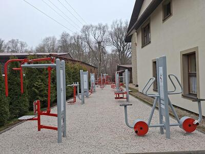 Urządzenia siłowni zewnętrznej na kamienistym podłożu, po bokach krzewy i ściana szkoły