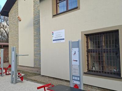 Trzy urządzenia siłowni zewnętrznej na tle ściany budynku szkoły z tablicą informacyjną