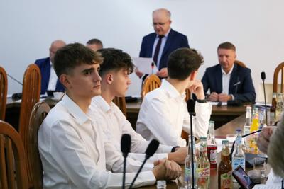 Przedstawiciele Młodzieżowej Rady Powiatu Myślenickiego. Trzech uczniów ubranych w białe koszule, siedzących przy stole z Radnymi.