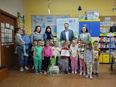 Grupa przedszkolaków i organizatorzy konkursu „Przedszkolaki w dobrym klimacie” z nagrodami. W tle baner projektu i kolorowe dekoracje.