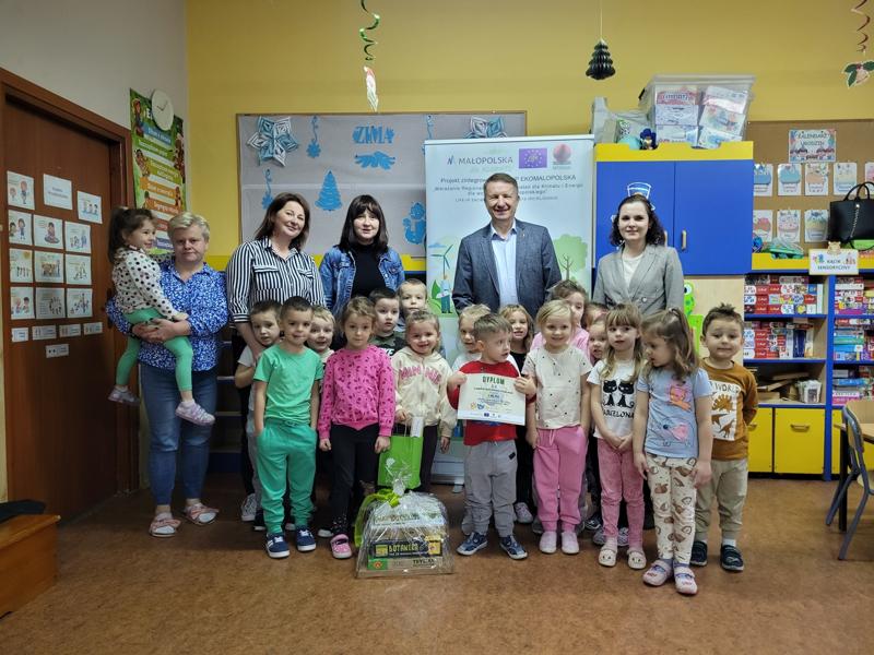 Grupa przedszkolaków i organizatorzy konkursu „Przedszkolaki w dobrym klimacie” z nagrodami. W tle baner projektu i kolorowe dekoracje.