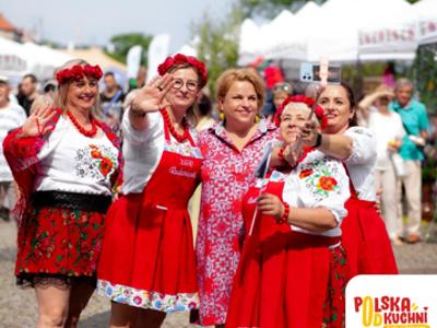 Na zdjęciu widoczne są kobiety w strojach regionalnych, które robią sobie zdjęcie. W dole widoczne jest logo.
