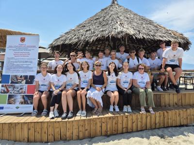 Grupa uczniów i nauczycielek w białych koszulkach z logo projektu Erasmus+ pozują na tle altany z trzcinowym dachem i baneru informacyjnego.