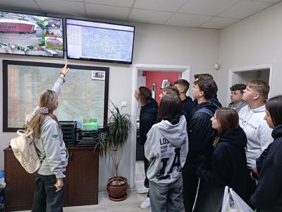 Grupa uczniów obserwuje prezentację prowadzoną przez kobietę wskazującą na ekran z mapą i monitoringiem w biurze.