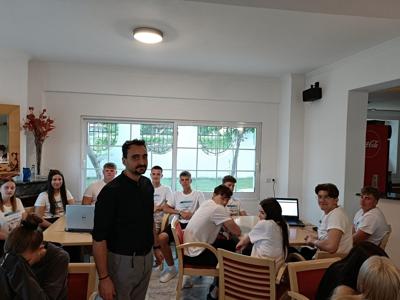 Grupa uczniów w białych koszulkach Erasmus+ siedzi przy stołach w jasnym pomieszczeniu z dużym oknem i widokiem na grecką zabudowę.