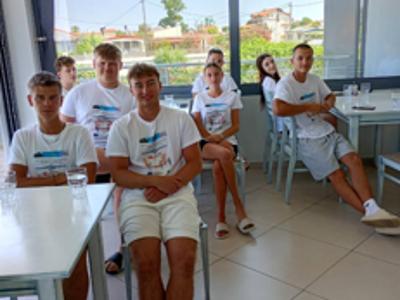 Nauczyciel prowadzi zajęcia dla uczniów ubranych w koszulki Erasmus+ – wszyscy siedzą przy stołach z laptopami.