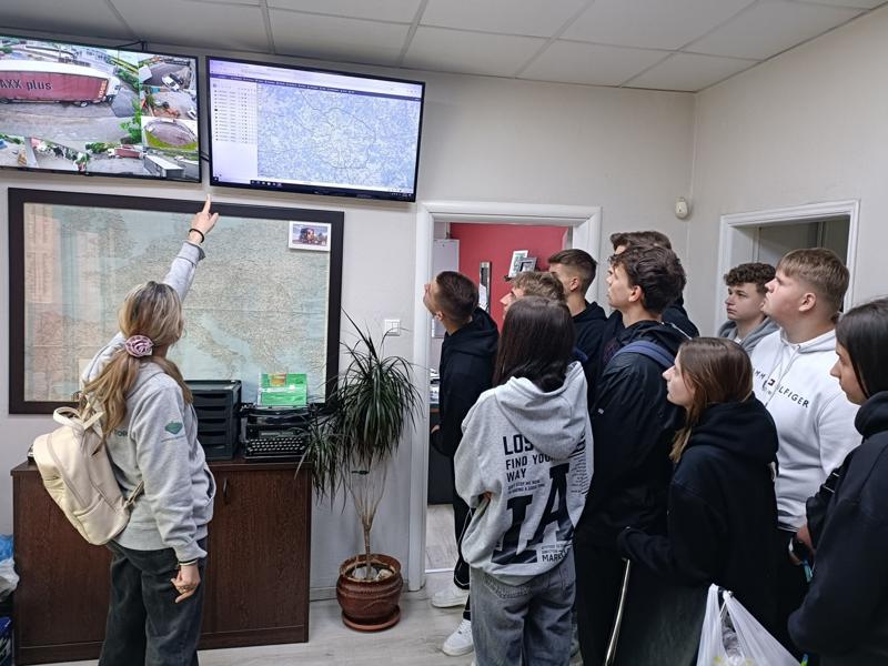 Grupa uczniów obserwuje prezentację prowadzoną przez kobietę wskazującą na ekran z mapą i monitoringiem w biurze.