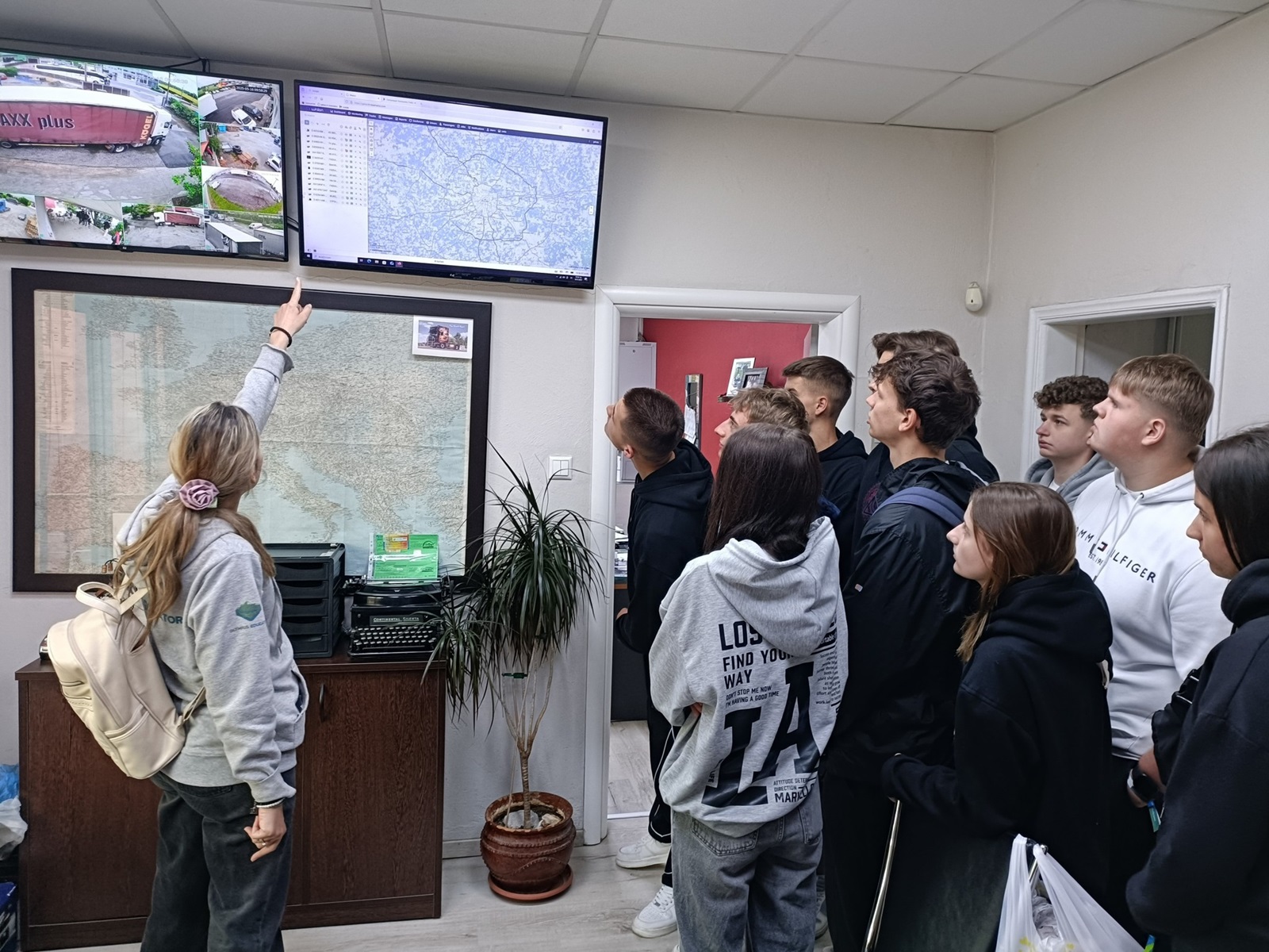 Grupa uczniów obserwuje prezentację prowadzoną przez kobietę wskazującą na ekran z mapą i monitoringiem w biurze.
