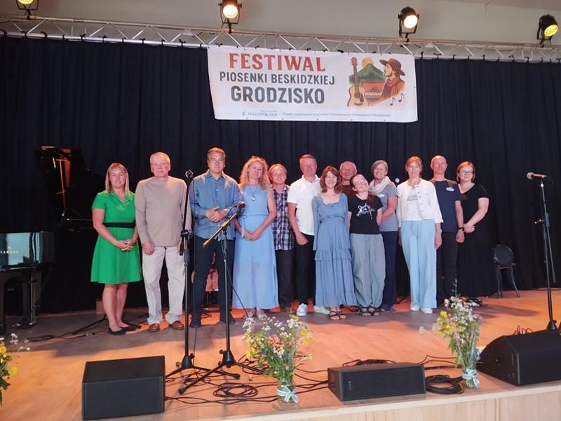 Grupa uczestników i organizatorów Festiwalu Piosenki Beskidzkiej w Grodzisku pozująca do wspólnego zdjęcia na scenie. W tle widoczny baner wydarzenia.