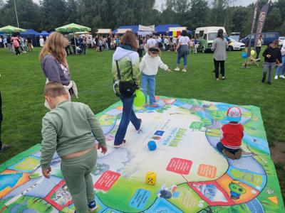 Na zdjęciu dzieci bawią się na dużej planszy edukacyjnej rozłożonej na trawie podczas plenerowego wydarzenia z udziałem rodzin.