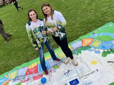Na zdjęciu dwie kobiety w zielonych bluzach z nadrukiem przyrody stoją na dużej edukacyjnej planszy rozłożonej na trawie podczas wydarzenia plenerowego.