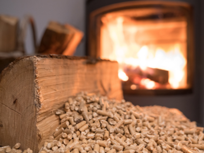 Zbliżenie na drewno opałowe i pellet drzewny przed kominkiem z płonącym ogniem w tle.