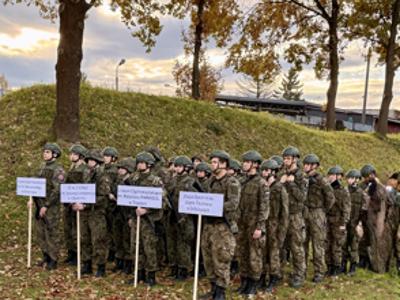 Grupa uczniów w mundurach podczas zawodów, trzymająca tabliczki z nazwami szkół o profilu wojskowym.