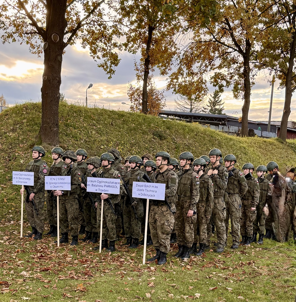 Grupa uczniów w mundurach podczas zawodów, trzymająca tabliczki z nazwami szkół o profilu wojskowym.