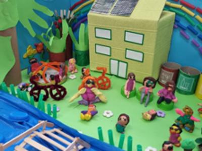 Praca przedstawiająca domek, rzekę, drzewa, łąkę i dzieci z surowców wtórnych i plasteliny