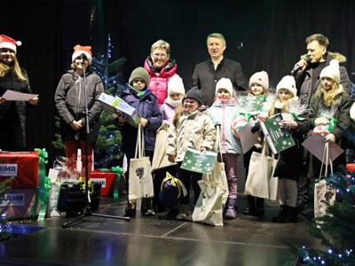 Czarna scena ze świątecznymi dekoracjami, na niej starości, dwie kobiety w czapkach mikołajowych, konferansjer i 8 dzieci trzymających nagrody