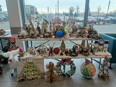 Wystawa ozdób świątecznych wykonanych z materiałów ekologicznych i naturalnych, ustawionych na stole i ławce, w tle za szybą parking z samochodami 