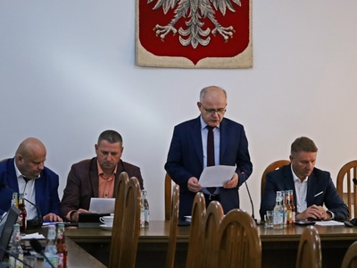 Starosta Józef Tomal oraz trzech Radnych siedzą przy stole. Przewodniczący Tadeusz Żaba stoi, czyta dokument. W tle herb Polski.