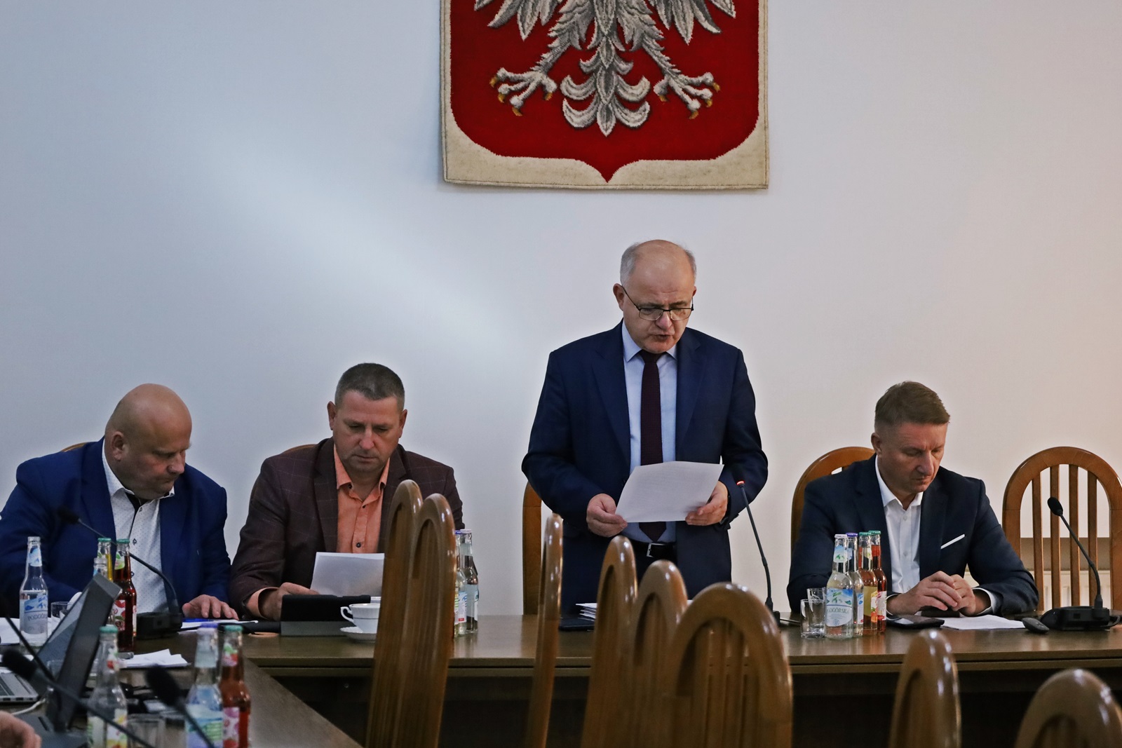 Starosta Józef Tomal oraz trzech Radnych siedzą przy stole. Przewodniczący Tadeusz Żaba stoi, czyta dokument. W tle herb Polski.