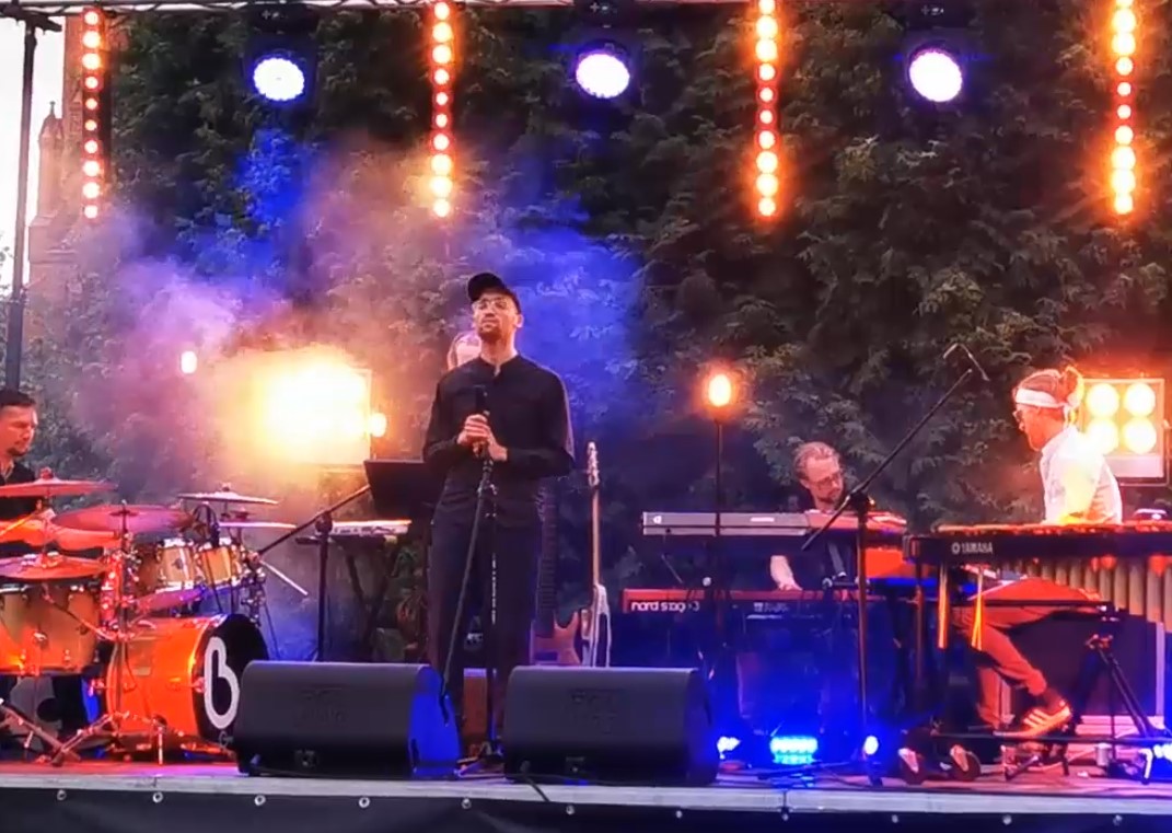 Kadr z filmu z koncertu przy katedrze. Na zdjęciu zespół na scenie i śpiewający wokalista.